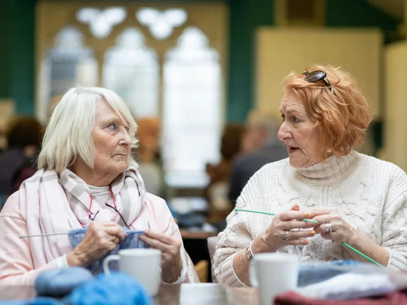 Two older women chatting over mugs of tea while knitting at a community group
